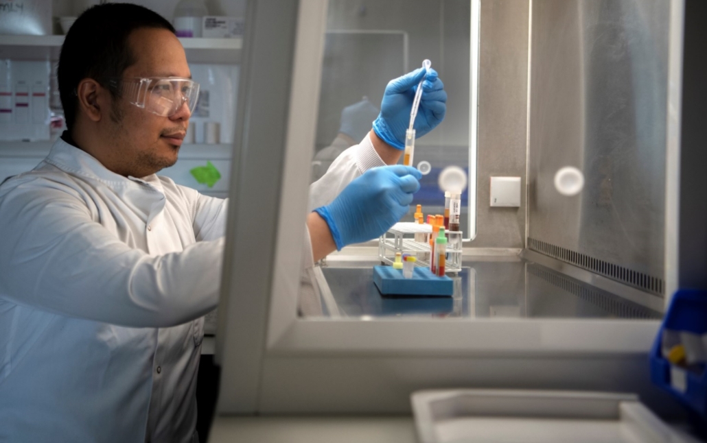 Member of staff in a white lab coat pipetting into a sample tube in a laboratory cabinet. Other samples are in a tube rack