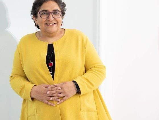 A lady with short dark hair with glasses, smiling wearing a yellow jacket